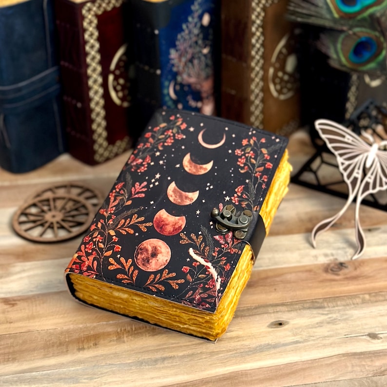 May include: A black leather journal with a moon and floral design. The journal has a brass clasp and is closed. The journal is sitting on a wooden surface.
