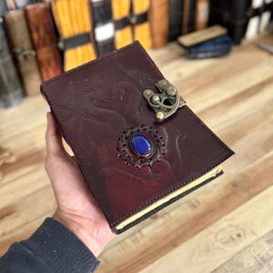May include: A burgundy leather journal with a dragon design and a metal clasp. The journal has a blue gemstone set in the center of the clasp.