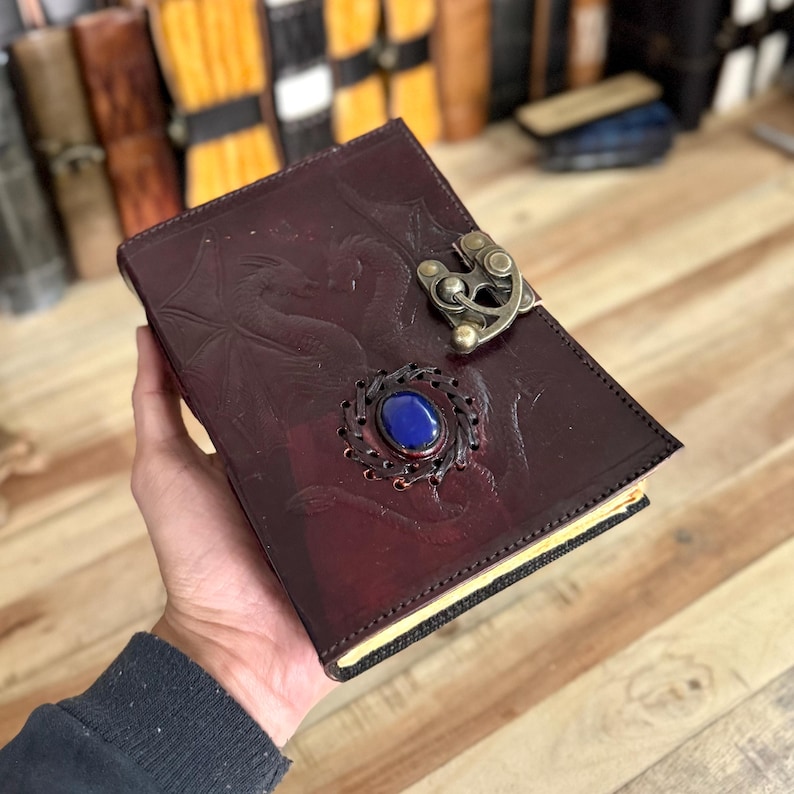 May include: A burgundy leather journal with a dragon design and a metal clasp. The journal has a blue gemstone set in the center of the clasp.