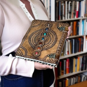 Puede incluir: Un diario de cuero hecho a mano con un dise&ntilde;o decorativo y piedras preciosas de colores. El diario presenta un patr&oacute;n en relieve detallado y un cierre de metal. Las piedras preciosas son rojas, naranjas, amarillas, verdes, azules y moradas.