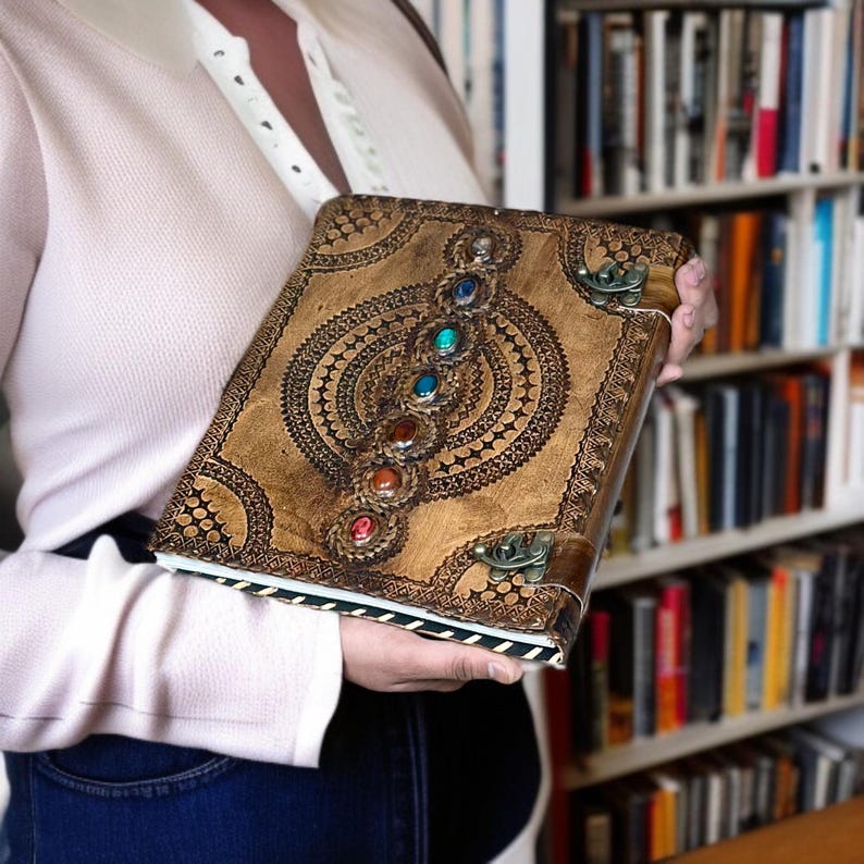 Puede incluir: Un diario de cuero hecho a mano con un dise&ntilde;o decorativo y piedras preciosas de colores. El diario presenta un patr&oacute;n en relieve detallado y un cierre de metal. Las piedras preciosas son rojas, naranjas, amarillas, verdes, azules y moradas.