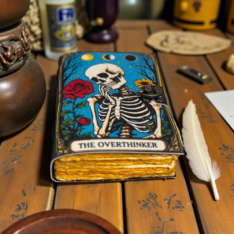 May include: A journal with a cover featuring a skeleton in thought, a red rose, and the text "THE OVERTHINKER" against a blue background. The journal has aged, yellowed pages and a metal clasp.