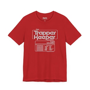 May include: Red t-shirt with a white graphic design. The design features the words "Trapper Keeper Notebook" in a retro font, along with a table of contents and useful information. The shirt is a classic crew neck style.