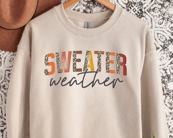 May include: A beige sweatshirt with the words "SWEATER weather" printed on the front. The word "SWEATER" is in a mix of colors and patterns, including leopard print, orange, and brown. The word "weather" is in a cursive font.