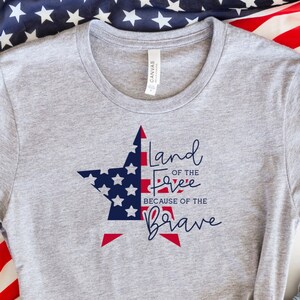 Puede incluir: Una camiseta gris con un diseño de estrella en rojo, blanco y azul y el texto "Land of the Free, Because of the Brave". La camiseta se combina con pantalones cortos de mezclilla y zapatillas blancas. Una bandera estadounidense es visible en el fondo, creando un tema patriótico.