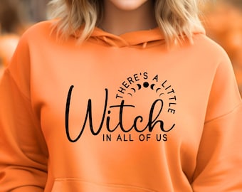 Op de afbeelding: Een oranje hoodie met de tekst "There's a little Witch in all of us" in het zwart. De hoodie heeft een capuchon met trekkoord en een voorzak. De persoon op de foto draagt een zwarte hoed en staat voor een pompoenveld.