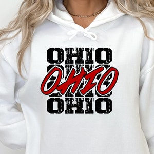 May include: White hoodie with a front pocket and drawstring. The design features the word "OHIO" in a distressed black font, with a red "OHIO" in the center. The hoodie is a casual style.