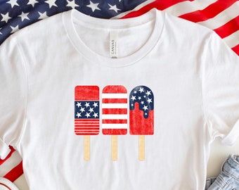 Könnte beinhalten: Weißes T-Shirt mit drei Eis am Stiel-Designs in Rot, Weiß und Blau, die an die amerikanische Flagge erinnern. Das Shirt wird mit Jeansshorts und weißen Turnschuhen kombiniert, vor dem Hintergrund der amerikanischen Flagge.