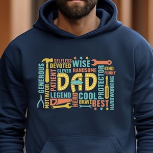 以下が含まれることがあります： 中央に「DAD」の文字、周りに「賢い」「クール」「ヒーロー」などの言葉が書かれた、ネイビーブルーのパーカー。デザインにはツールと星が組み込まれています。フロントポケットが見えます。