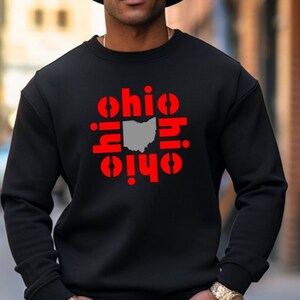 May include: Black crewneck sweatshirt with the word "Ohio" in red, surrounding a gray outline of the state. The person in the image is wearing a black cowboy hat and blue jeans.