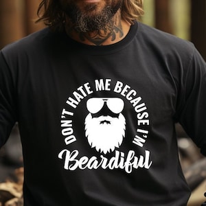 Puede incluir: Camiseta negra de manga larga con el texto blanco "DON'T HATE ME BECAUSE I'M BEARDIFUL". El gráfico presenta una barba blanca y gafas de sol. La persona de la imagen lleva un gorro negro y un reloj.
