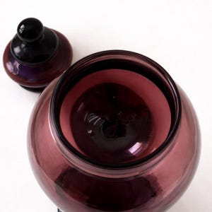 May include: A clear, round, amethyst-colored glass jar with a matching lid. The jar has a wide opening and sits on a black base. The lid is also black with a small, round handle. The jar is empty.
