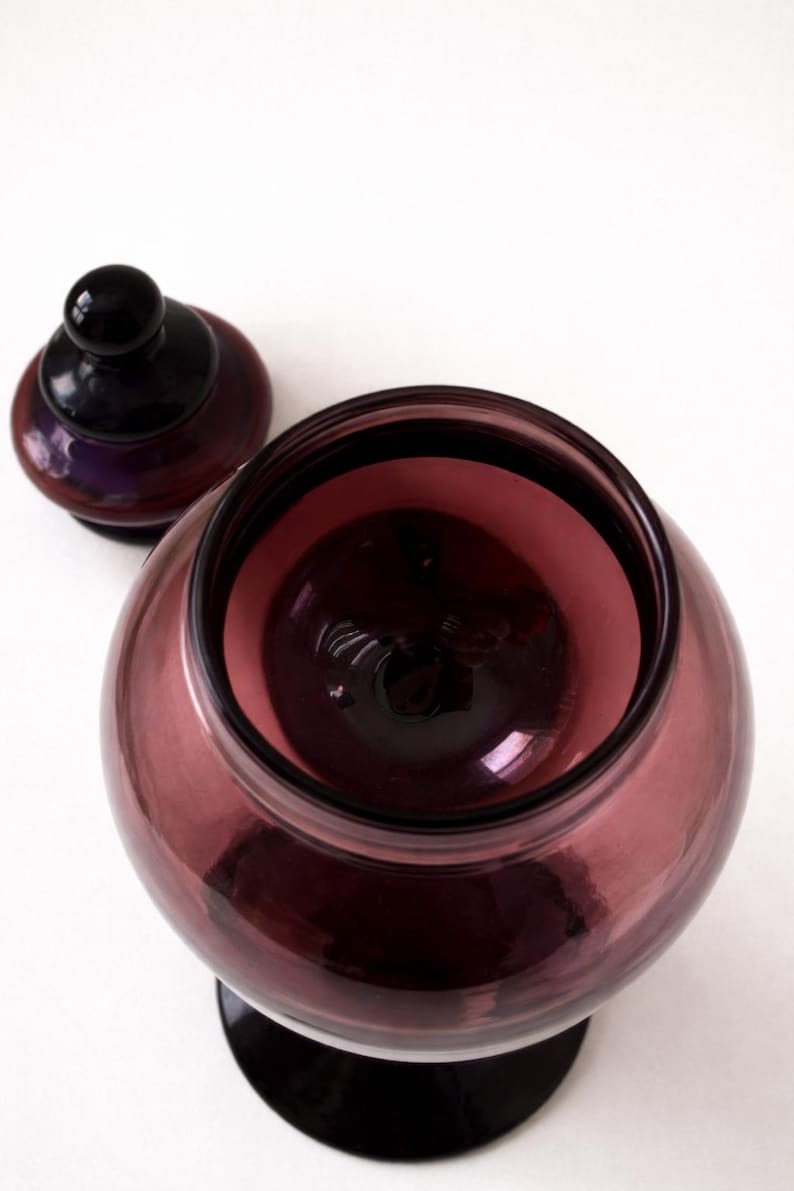 May include: A clear, round, amethyst-colored glass jar with a matching lid. The jar has a wide opening and sits on a black base. The lid is also black with a small, round handle. The jar is empty.