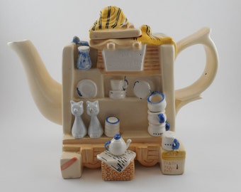 Op de afbeelding: Een keramische theepot in de vorm van een kleine winkel met een geel gestreept luifel, twee witte katten en een stapel blauwe en witte theekopjes. De theepot heeft een blauw en wit theekopje aan de zijkant en de tekst "CAPRI" en "FINEST TEA POT" aan de onderkant.