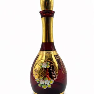 Puede incluir: Una jarra de cristal rojo rubí con detalles dorados y un tapón a juego. La jarra presenta diseños florales y de vid pintados en oro. El cuello y la base también están adornados con oro. Una pieza decorativa para el hogar.