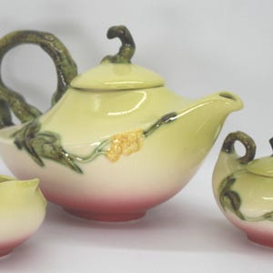 Op de afbeelding: Een keramische theeset met een theepot, melkkan en suikerpot. De set heeft een lichtgele en roze kleurstelling met groene wijnranken en gele bloemaccenten. De theepot heeft een grote, gebogen handgreep.