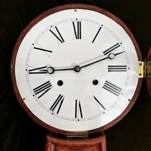 May include: A round wall clock with a white face, black Roman numerals, and two thin black metal hands. The clock features a dark wood frame, two circular winding holes, and a brass hinge on the right side connecting to a lower wooden section.