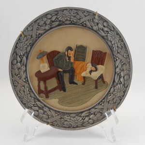 May include: Decorative plate with a raised relief scene of a man sitting near a sleeping person. The plate has a silver-colored floral border and is displayed on a clear stand. The scene depicts a room with chairs and a table.