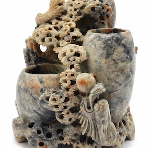 Puede incluir: Escultura de piedra tallada con tres vasijas y detalles intrincados. La escultura tiene una paleta de colores neutros con tonos de gris, beige y marrón. El diseño incluye formas florales y orgánicas, con una superficie texturizada.