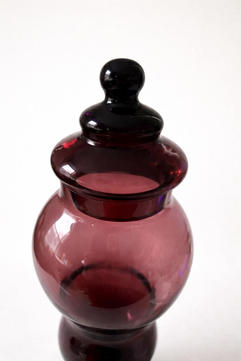 May include: A decorative glass jar with a rounded body and a lid. The jar is a deep purple color, with a glossy finish. The lid is a darker shade of purple. The jar is likely used for storage or display.