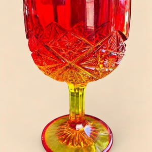 May include: A vibrant red and yellow ombre glass goblet featuring a scalloped rim and a detailed diamond-cut pattern across the bowl. The stem is faceted, leading down to a circular base with a sunburst design radiating from the center point.