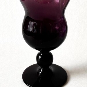 May include: A tall, elegant wine glass with a deep purple hue. The glass features a rounded bowl, a slender stem with a spherical detail, and a wide, circular base. The glass is made of a glossy material, reflecting light.