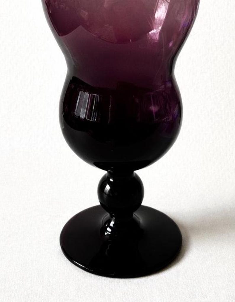 May include: A tall, elegant wine glass with a deep purple hue. The glass features a rounded bowl, a slender stem with a spherical detail, and a wide, circular base. The glass is made of a glossy material, reflecting light.