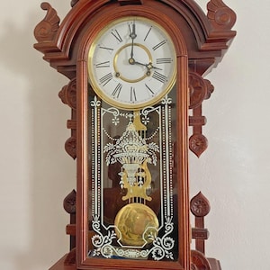 Puede incluir: Un reloj de pared de madera vintage con una esfera blanca y números romanos dorados. El reloj tiene un panel de vidrio decorativo con un diseño grabado y un péndulo. El reloj está hecho de madera marrón oscuro y tiene tallas ornamentadas.