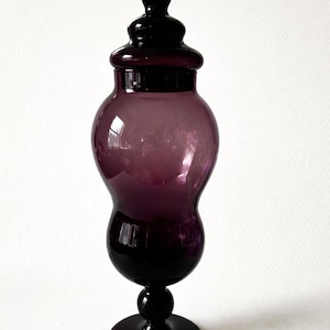 May include: A tall, elegant purple glass jar with a lid. The jar has a bulbous body and a slender base, and it stands on a round foot. The lid is also purple and has a small knob on top. The jar is approximately 12 inches tall.