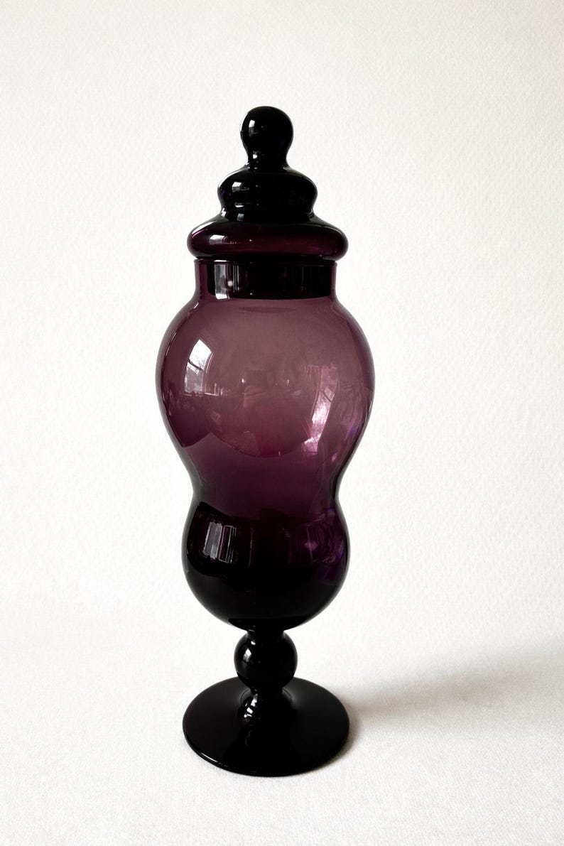 May include: A tall, elegant purple glass jar with a lid. The jar has a bulbous body and a slender base, and it stands on a round foot. The lid is also purple and has a small knob on top. The jar is approximately 12 inches tall.