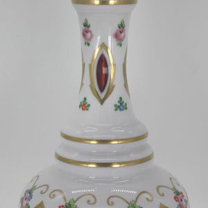 Puede incluir: Una jarra de vidrio blanco con un tapón decorativo. La jarra presenta adornos dorados y detalles ovalados rojos. Diseños florales en rosa, azul y verde adornan el cuerpo. La jarra mide aproximadamente 30 cm de altura.