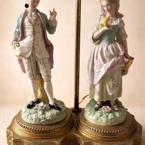Könnte beinhalten: Dekorative Lampe mit zwei Porzellanfiguren in historischer Kleidung. Die Figuren stehen auf verzierten Sockeln mit einem goldfarbenen Metallrahmen. Der Mann trägt einen hellblauen Mantel und die Frau hält einen gelben Gegenstand.