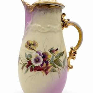 May include: Cream-colored porcelain pitcher with a lid, adorned with a floral design featuring purple, yellow, and burgundy pansies. The pitcher has a pink ombre effect and gold accents on the handle and lid.