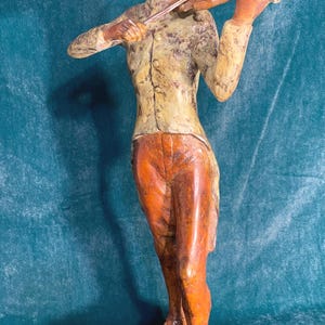 May include: A detailed sculpture of a person playing a violin. The figure is dressed in a yellow jacket and orange pants, standing on a black base. The violin is held up to the figure's chin. The sculpture has a vintage, antique appearance.