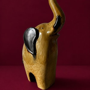 May include: A ceramic elephant figurine with a glossy tan and brown mottled finish. The elephant is depicted in a standing position with its trunk raised upward. It features distinct, smooth black ears and a small, dark eye detail on its face.