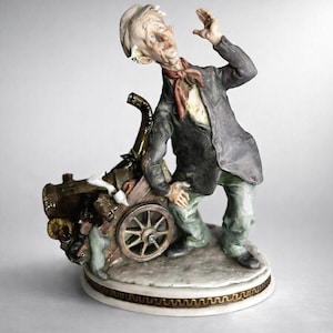 May include: A detailed porcelain figurine of an elderly man in a suit, leaning on a cart. The man has a surprised expression, a red scarf, and a white hat. The cart has a wooden wheel and is filled with various items. The figurine is on a round base.