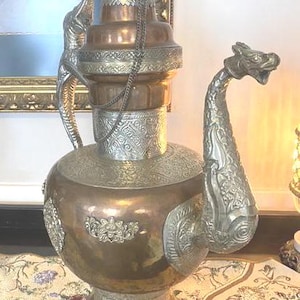 May include: An ornate copper and silver-toned metal pitcher with intricate detailing. The pitcher features a dragon-shaped spout and decorative patterns. A chain hangs from the top. The pitcher is likely a decorative item or a vessel for serving.