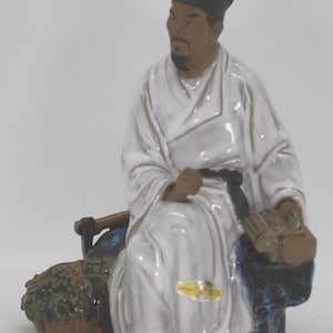 Puede incluir: Figura de cerámica de un hombre sentado con túnicas blancas y sombrero negro. Sostiene un libro y está sentado junto a una cesta de hojas verdes. La figura tiene un cinturón negro y está sobre una base azul y marrón.
