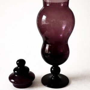 May include: A dark purple glass vase with a matching lid. The vase has a bulbous body and a flared top, resting on a round base. The lid is a small, rounded shape. The glass has a glossy finish.