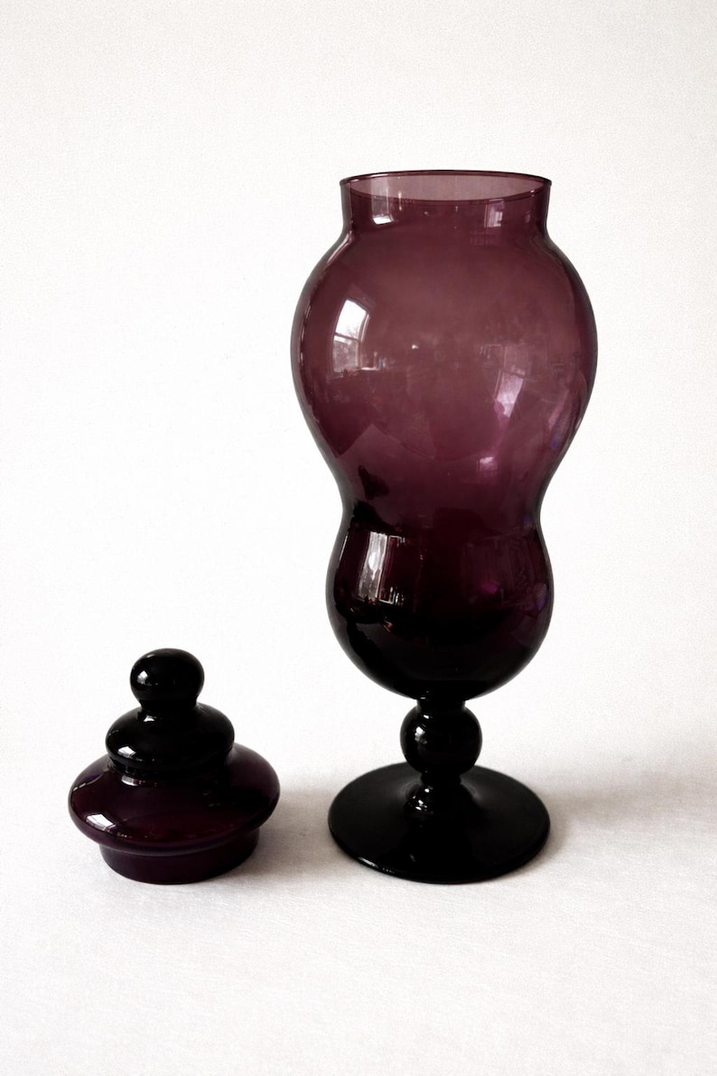 May include: A dark purple glass vase with a matching lid. The vase has a bulbous body and a flared top, resting on a round base. The lid is a small, rounded shape. The glass has a glossy finish.