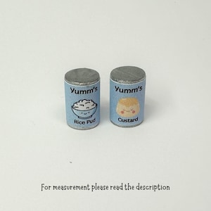 Könnte beinhalten: Zwei kleine, silberne Dosen mit blauen Etiketten. Die Etiketten zeigen "Yumm's" und ein Cartoon-Bild einer Schüssel mit Milchreis und einer Schüssel mit Pudding. Der Text auf den Etiketten lautet "Rice Pud" und "Custard".