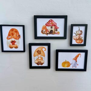 May include: Five framed watercolour art prints with black frames. The prints depict autumn-themed illustrations, including gnomes, pumpkins, and a mushroom house, in shades of orange, brown, and white. The artwork is displayed against a white wall.