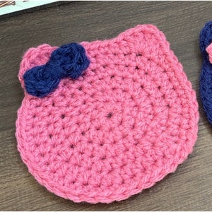 May include: A pink crocheted coaster with a blue yarn bow on top. The coaster is made with a simple stitch pattern and has a rounded shape.