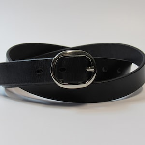 Puede incluir: Un cinturón de cuero negro con una hebilla ovalada plateada. El cinturón está enrollado sobre un fondo blanco. La hebilla es de color plateado brillante y el cinturón es de color negro liso.