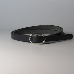 May include: A black leather belt with a silver oval buckle. The belt is thin and appears to be made of a smooth leather material. The buckle is a simple, classic design.