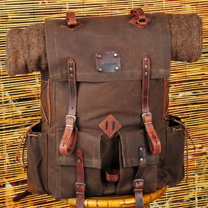May include: A brown canvas backpack with leather straps and buckles. The backpack has a large main compartment, two front pockets, and a small pocket on the top. The backpack is sitting on a wooden stand.