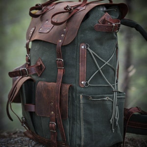 May include: A green canvas and brown leather backpack with multiple pockets and straps. The backpack has a drawstring closure and a flap with a buckle.