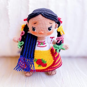 A crochet doll is dressed in clothing indigenous to Michoacán. Her black hair is in two braids with colorful ribbons. She wears an embroidered blouse and gold apron and a striped skirt. A blue and black rebozo hangs over her shoulder.