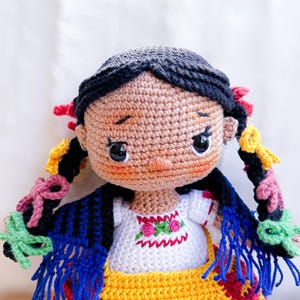 A close up of the face of a crochet doll wearing Purépecha clothing. Her black hair is in two braids lined with colorful bows. She is wearing a blue and black rebozo over her embroidered blouse.