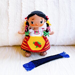 A crochet doll wears Purépecha clothing, including an embroidered white blouse and gold apron, a striped skirt and brown sandals. Her black hair is in two braids adorned with colorful ribbons. A blue and black rebozo lays spread out in front of her.